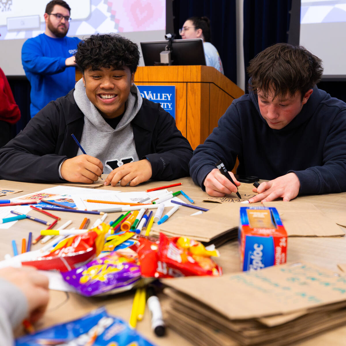 Two students sitting at a table and coloring on brown paper bags